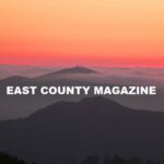 East County Magazine logo - text "east county magazine" in white; background image of sunset over mountains.