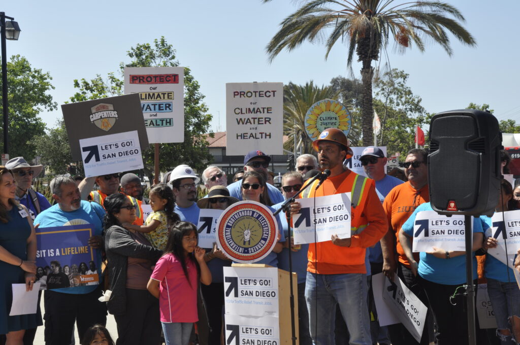 A person speaking in a microphone in front of people holding up signs about getting better transit in San Dieg.