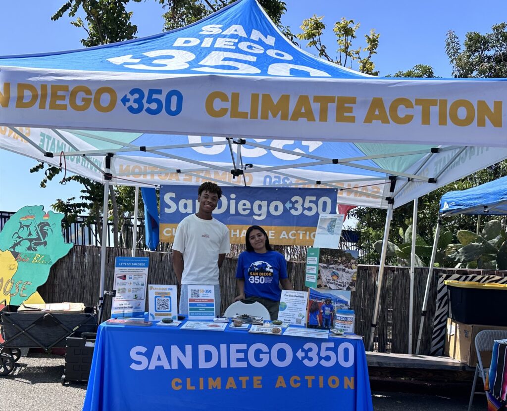 Two SD350 fellows smiling under a SD350 canopy and behind a table that reads SanDIego350. 