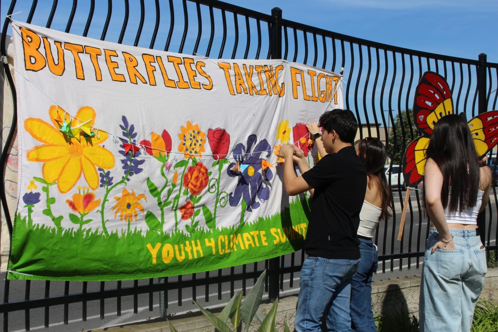 banner on a fence that reads "butterflies taking flight" with youth adding flowers to the banner